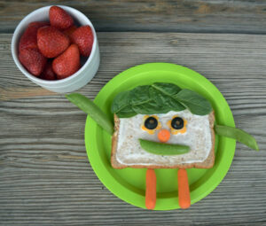 A sandwich has vegetable face and vegetables with a face on plate, strawberries in bowl.